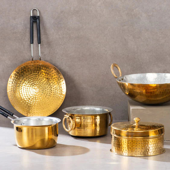 A collection of hammered brass cookware including pots, a bowl, and a frying pan against a neutral background.